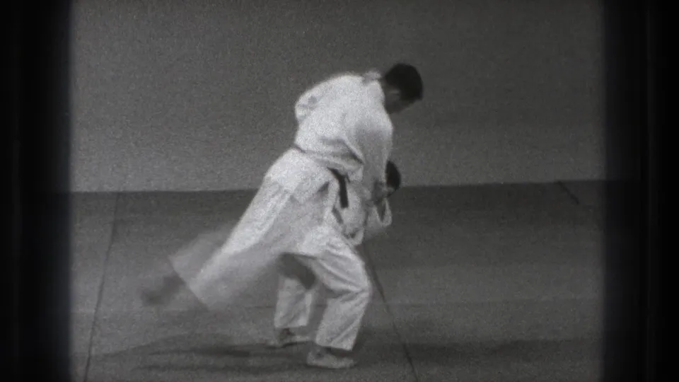 In 1971 Tokyo footage, two adult martial artists in white gi engage at the start of a judo-style throw on a dojo mat.