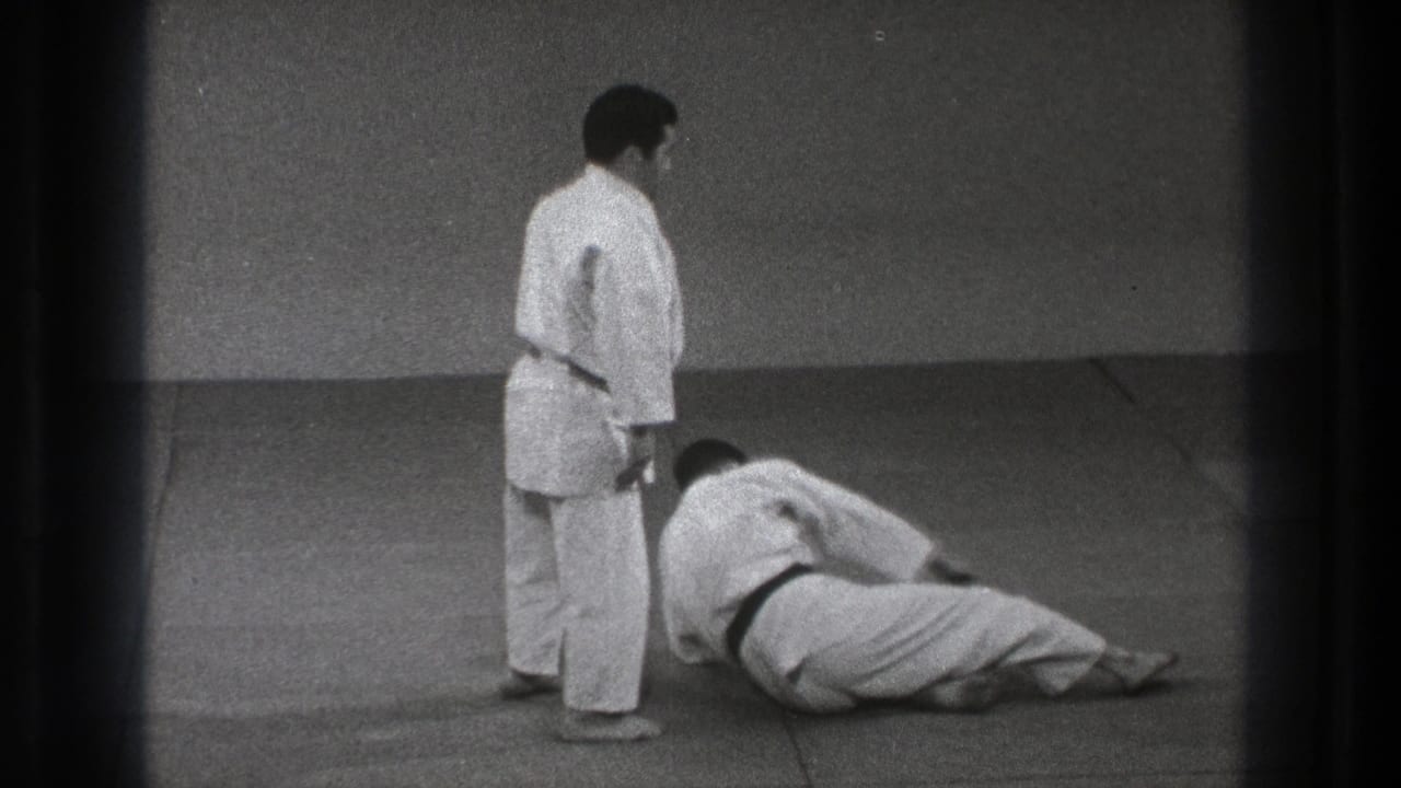 After the throw, one adult martial artist stands beside the other, who remains seated or reclining on the dojo mat in 1971 Tokyo footage.