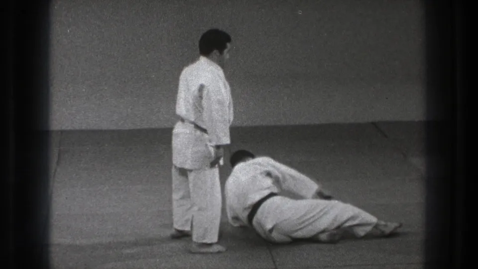 After the throw, one adult martial artist stands beside the other, who remains seated or reclining on the dojo mat in 1971 Tokyo footage.