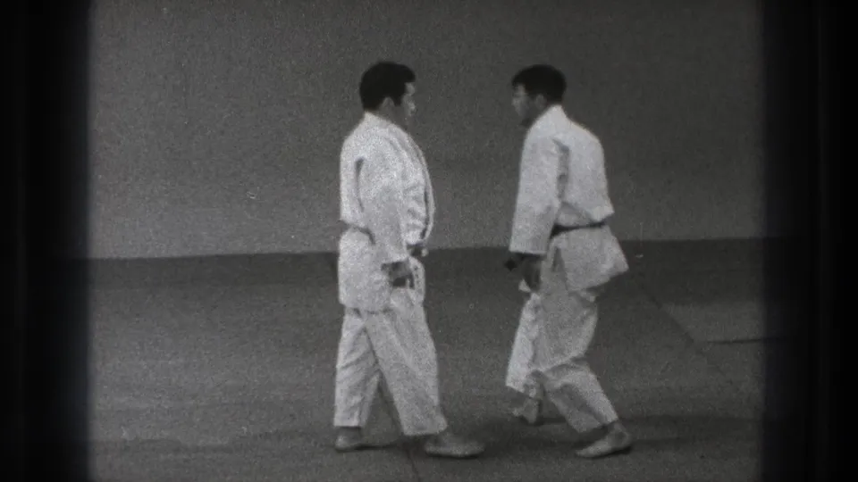 Two adult karate practitioners in white gi face each other in a sparse Tokyo dojo setting from the 1971 home-movie clip.