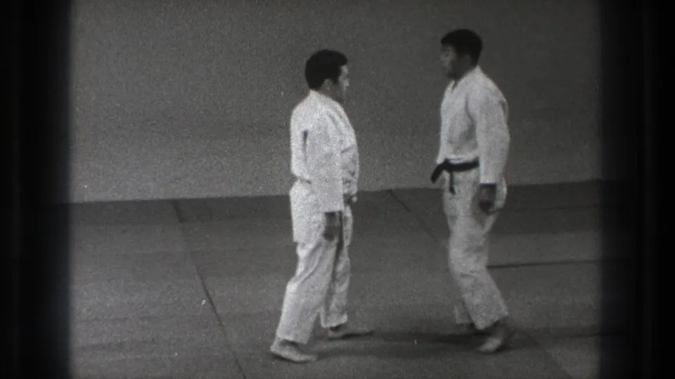 In the 1971 Tokyo dojo footage, a black-belt practitioner faces another man in a white gi on the training mat.