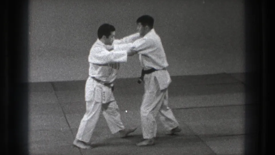 In 1971 Tokyo footage, two men in white gi face each other in a paired martial arts grip on a mat.