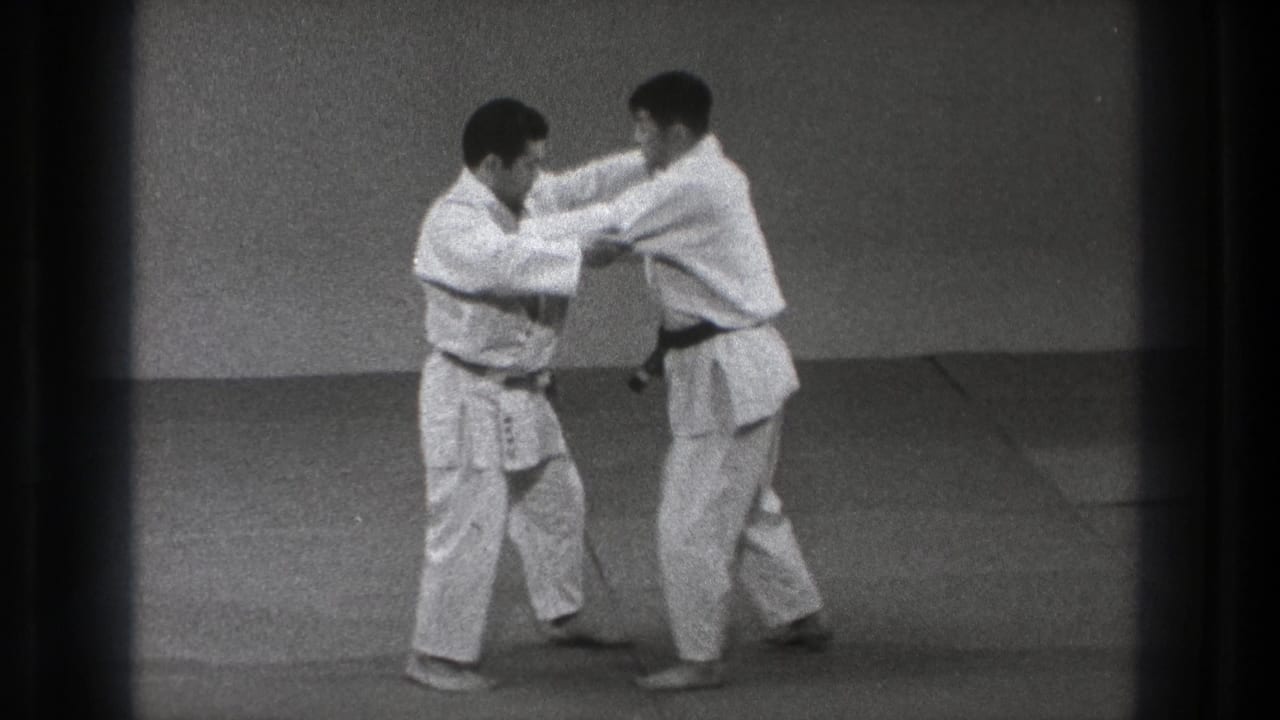Two men in gi continue a close paired technique on a dojo mat in the 1971 Tokyo home-movie clip.