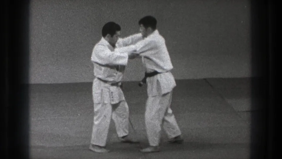Two men in gi continue a close paired technique on a dojo mat in the 1971 Tokyo home-movie clip.