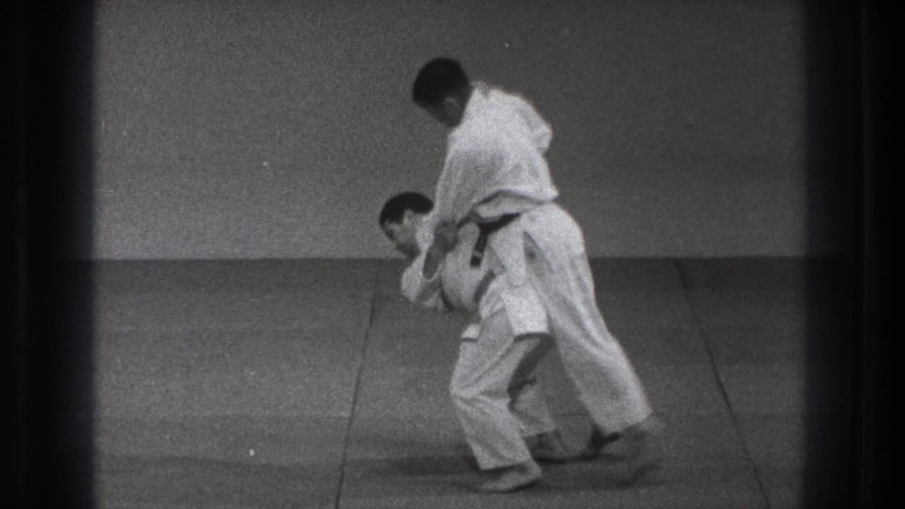 The paired technique reaches a bent-forward position, with one man controlling the other on the dojo mat.