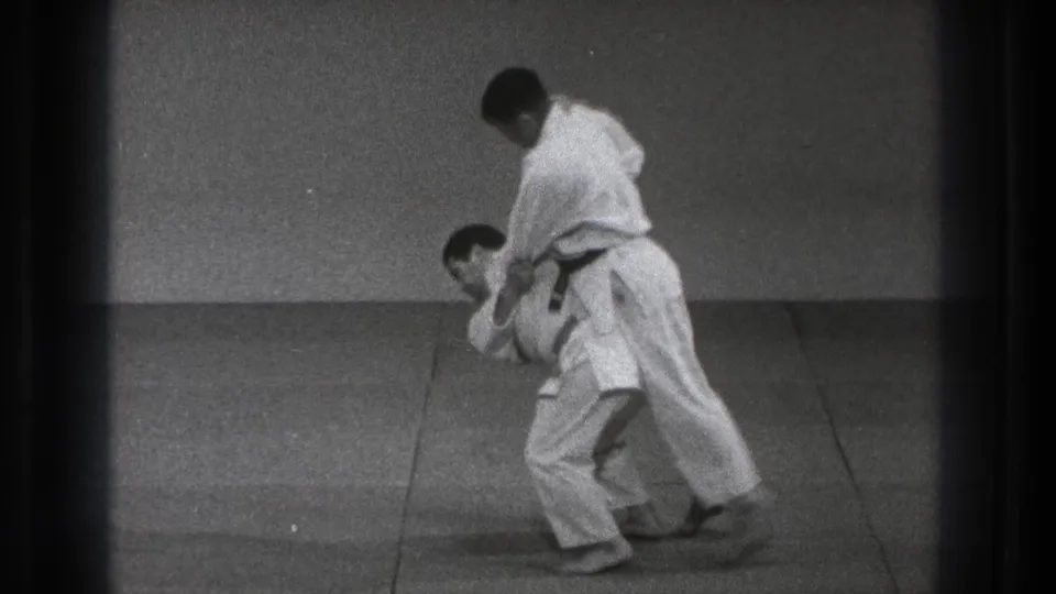 The paired technique reaches a bent-forward position, with one man controlling the other on the dojo mat.