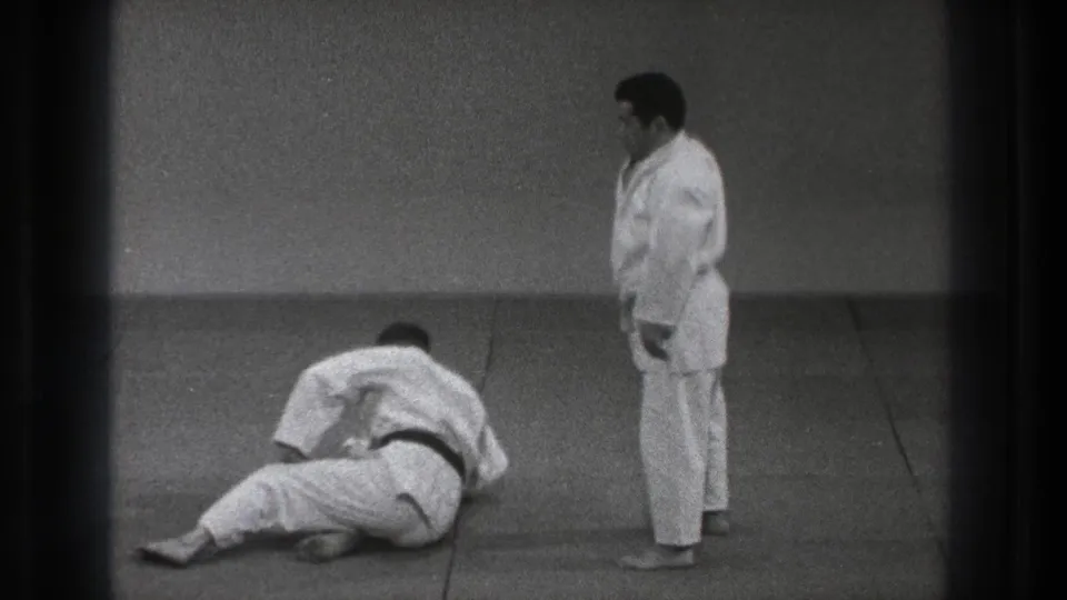 The thrown partner remains on the mat while the other martial artist stands beside him in the training room.