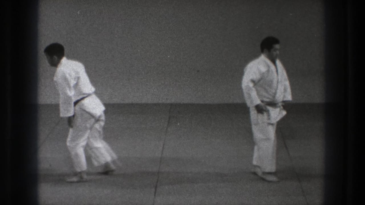 Two men in white martial arts uniforms stand on a grey mat in a plain indoor room.