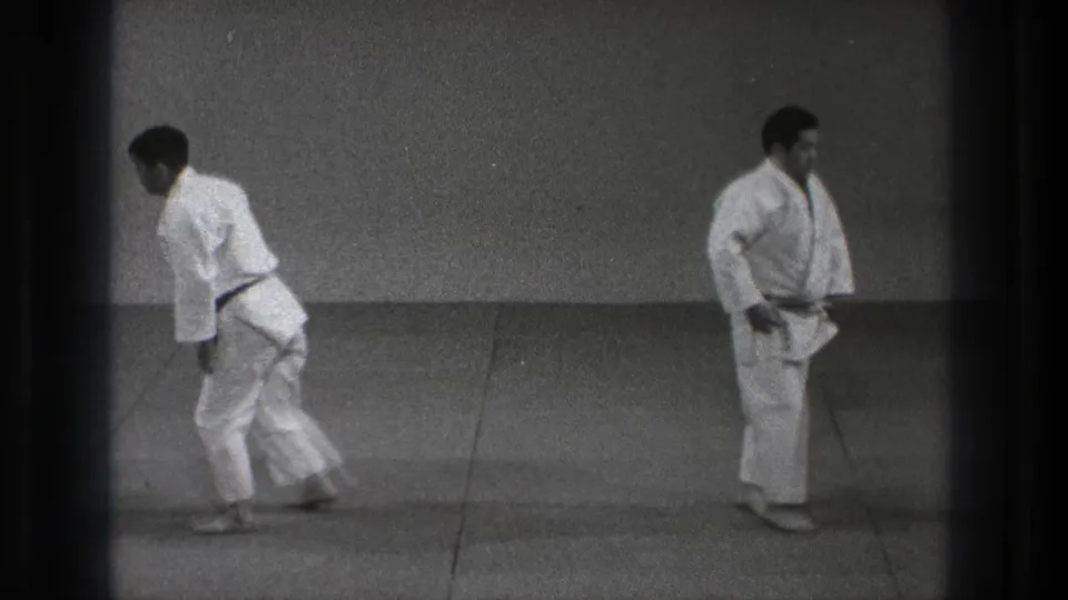 In Tokyo 1971 footage, two adult men in white karate uniforms are spaced apart on a grey mat in a simple dojo-like room.