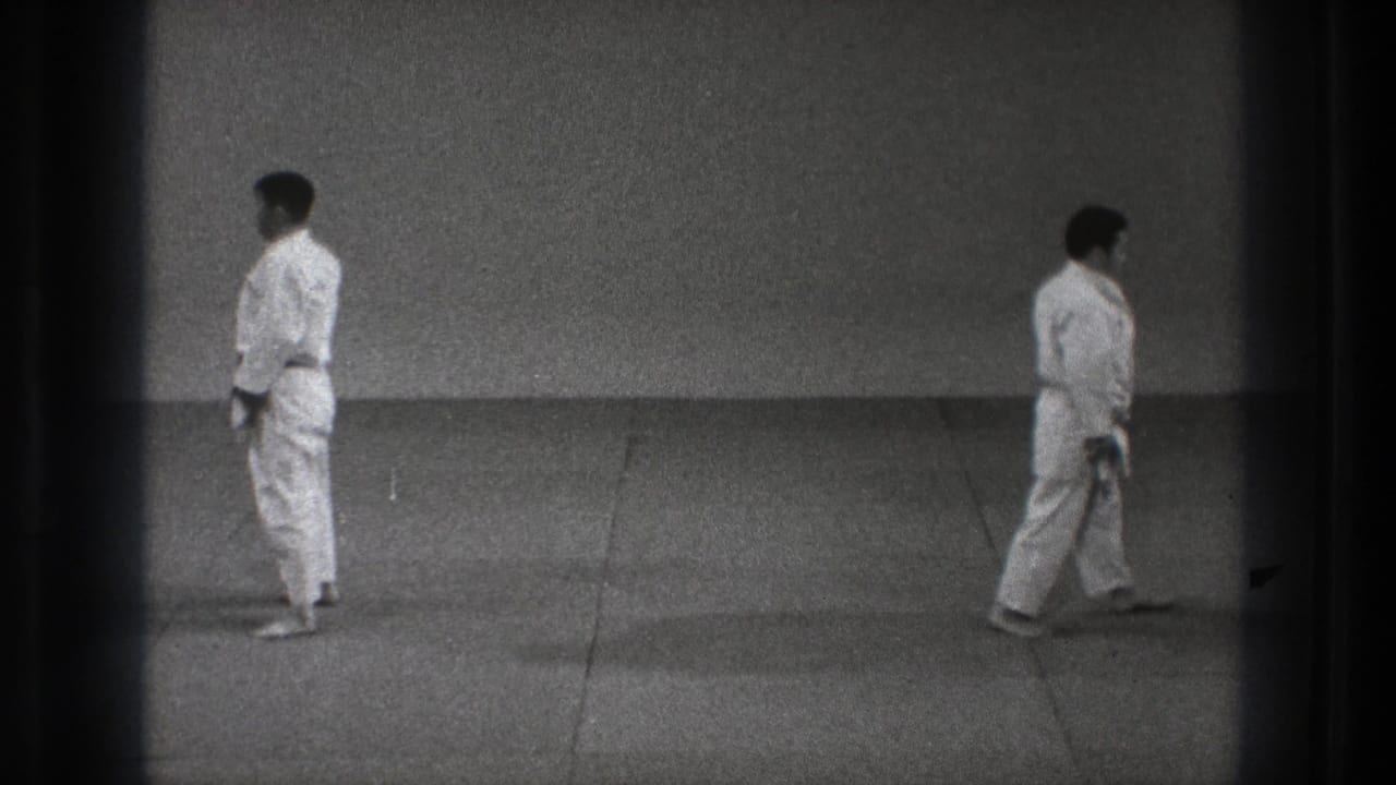 Two adult men in white karate uniforms stand at opposite sides of the mat in the Tokyo 1971 indoor training scene.