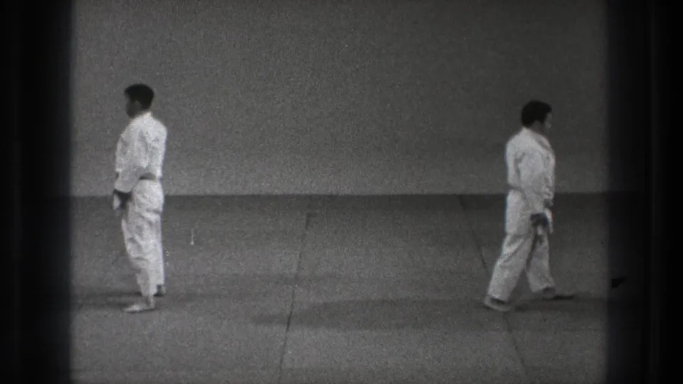 Two adult men in white karate uniforms stand at opposite sides of the mat in the Tokyo 1971 indoor training scene.