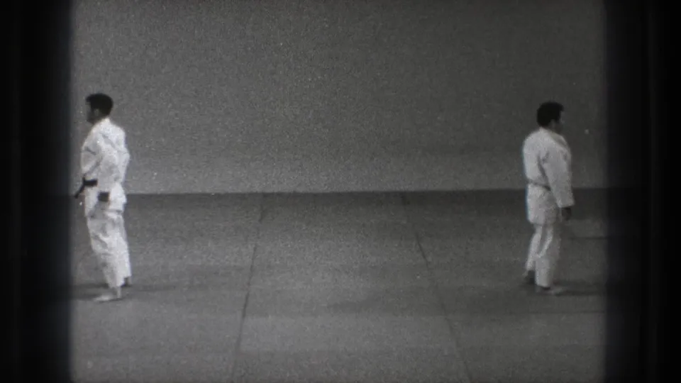 A Tokyo 1971 8mm frame shows one uniformed man near the left edge and another crossing the grey dojo mat.