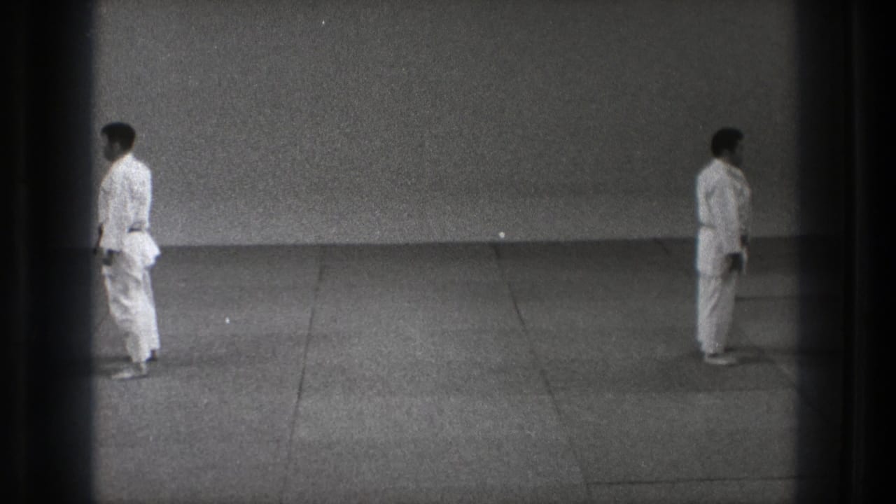 Two men in white karate gi stand apart on a plain mat in black-and-white footage.