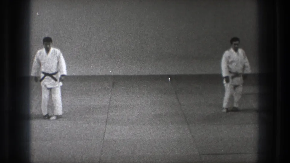 1971 Tokyo 8mm frame showing two adult martial artists in white gi and black belts facing on a plain dojo mat.