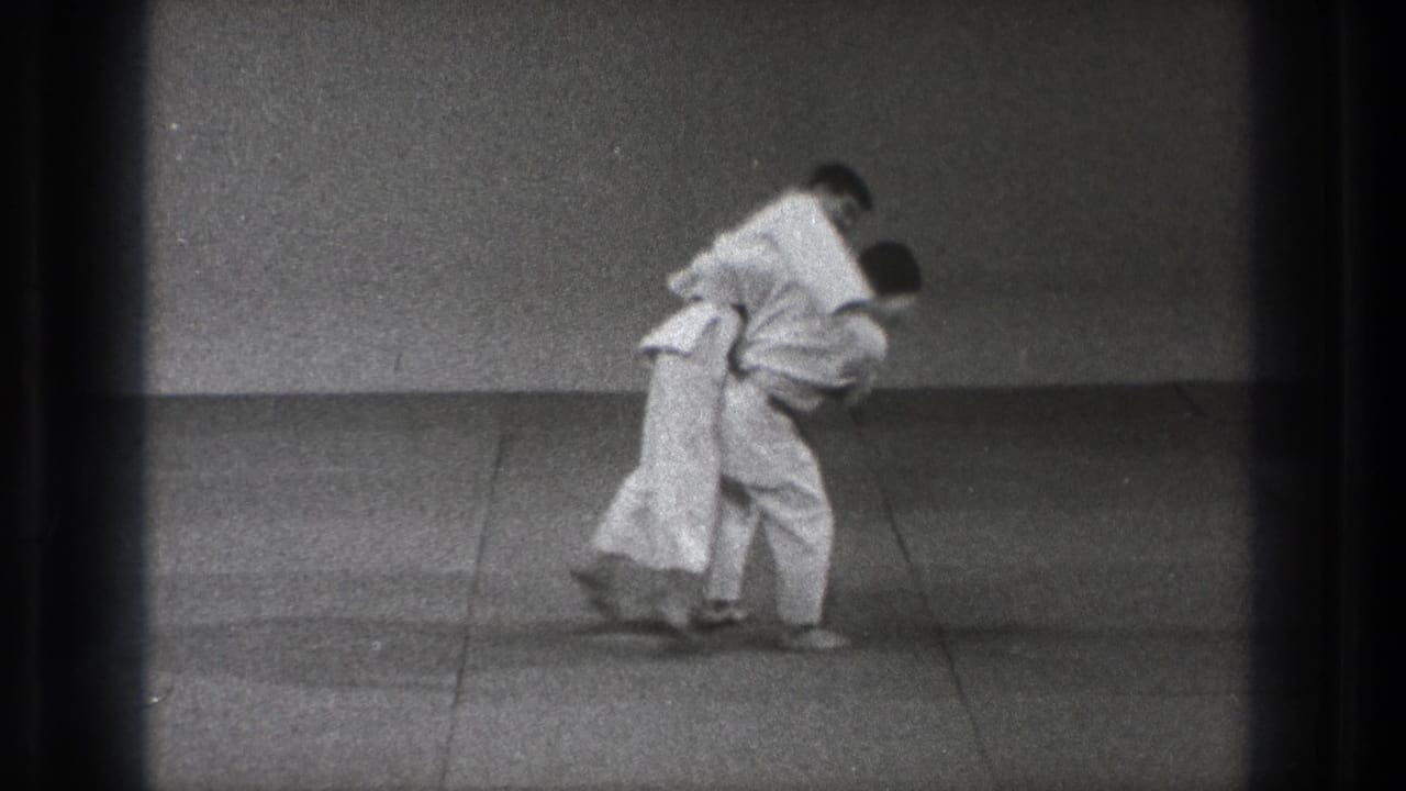The judo pair bend forward during the throw, with one man drawing the other across his hip on the mat.