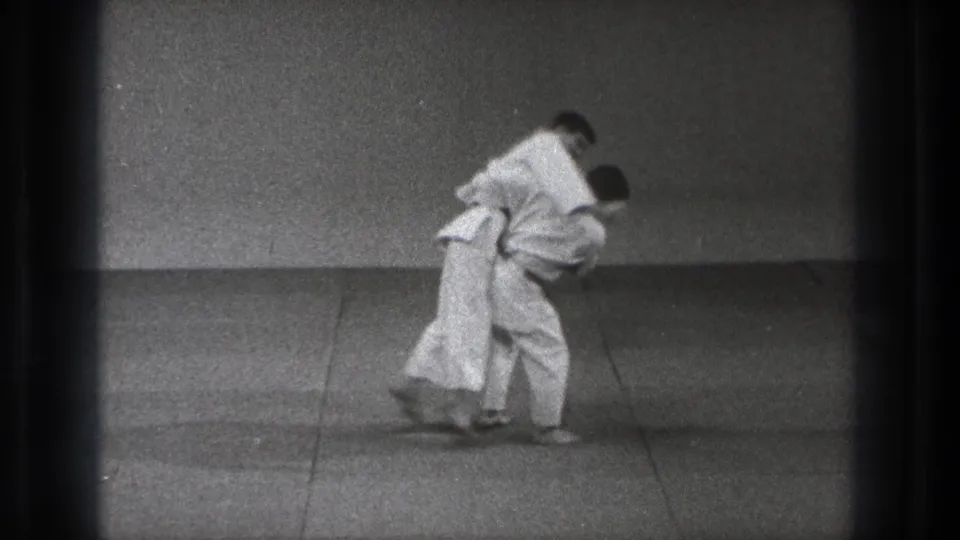 The judo pair bend forward during the throw, with one man drawing the other across his hip on the mat.