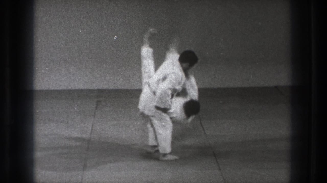 Two men in white gi practice a throw on a grey indoor mat.