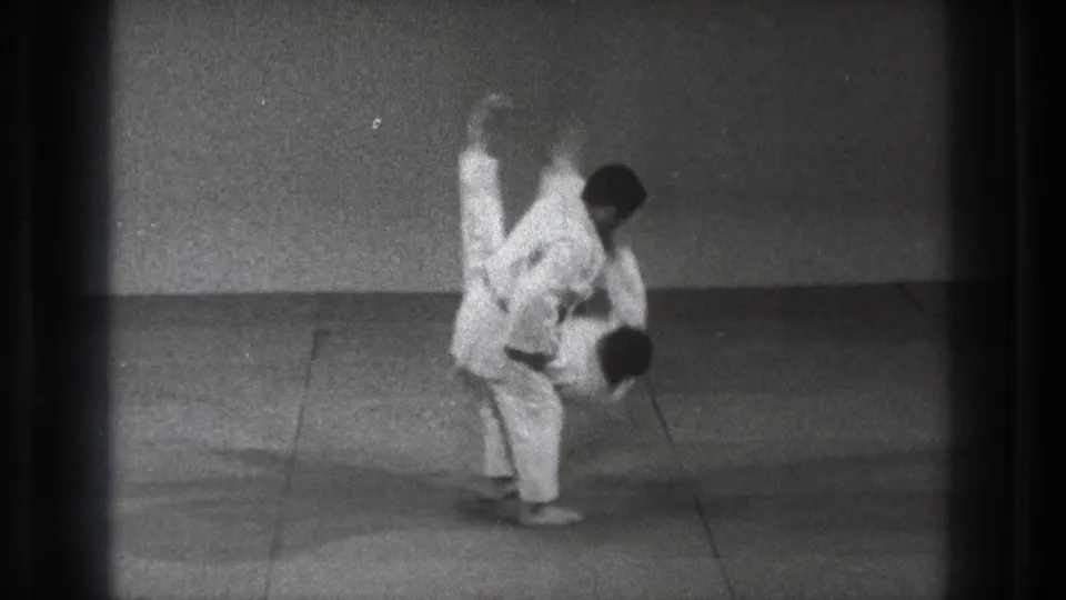 In 1971 Tokyo footage, one man in a white gi lifts another during a martial arts throw on a plain grey mat.