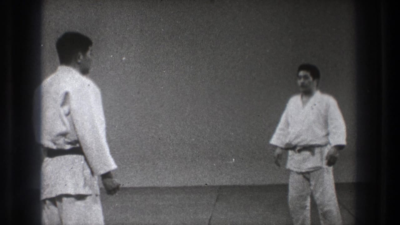 Two adult men in white martial arts uniforms stand on a grey mat in a plain Tokyo indoor practice room.