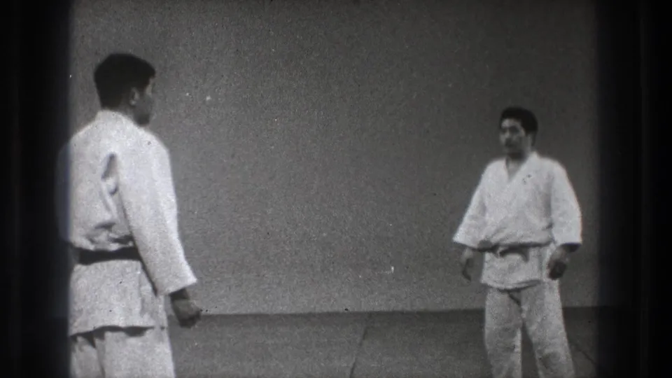 Two adult men in white martial arts uniforms stand on a grey mat in a plain Tokyo indoor practice room.