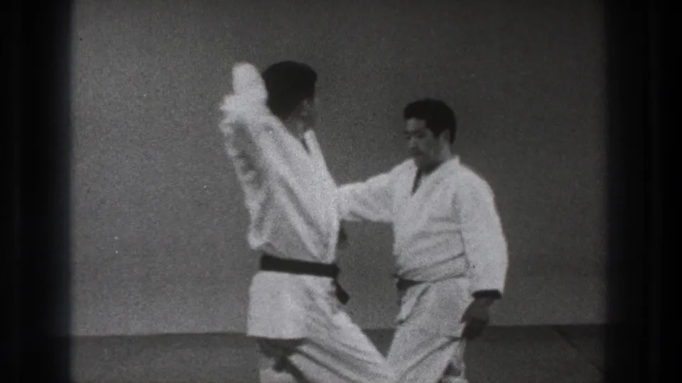 One man raises an arm while the other reaches in during a close martial arts training exchange in 1971 Tokyo footage.