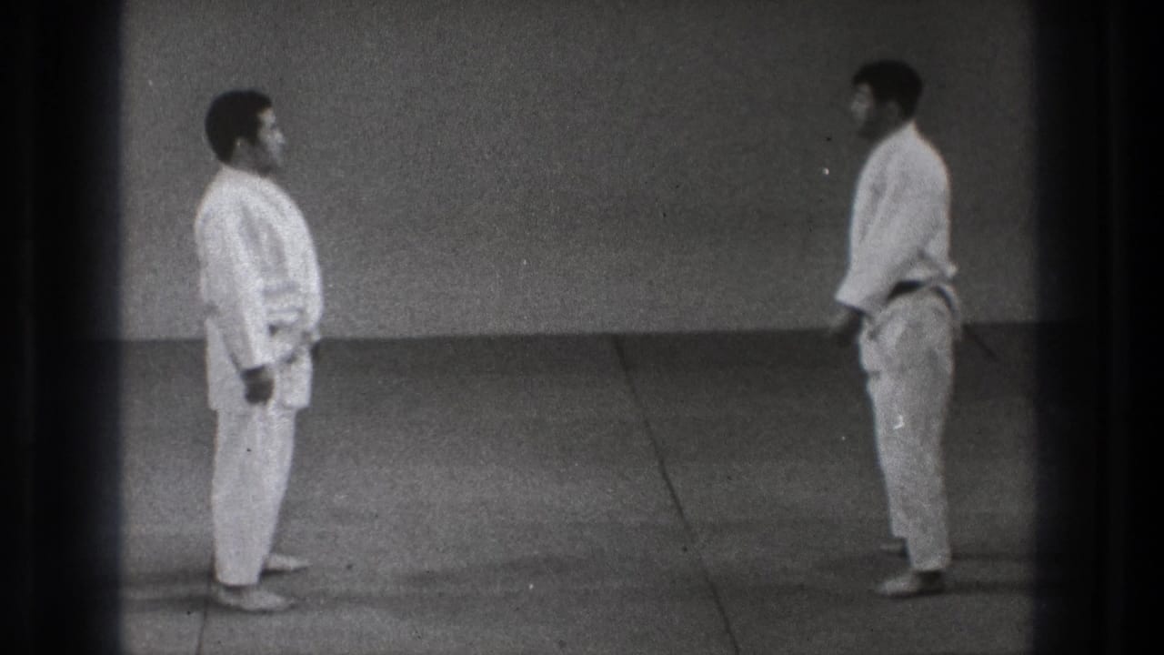 Two martial arts practitioners in white gi stand apart facing each other in the Tokyo 1971 practice space after the throwing sequence.