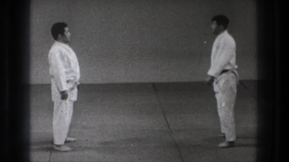 Two martial arts practitioners in white gi stand apart facing each other in the Tokyo 1971 practice space after the throwing sequence.