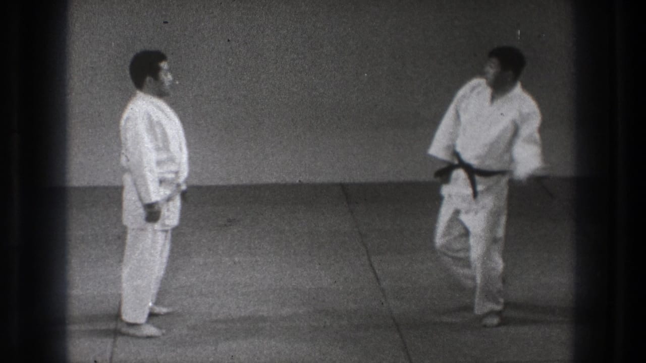 In 1971 Tokyo footage, two men in white gi stand on a dojo mat while a black-belt participant starts a technique.