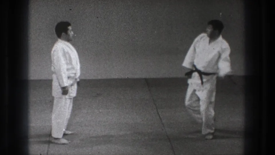 In 1971 Tokyo footage, two men in white gi stand on a dojo mat while a black-belt participant starts a technique.