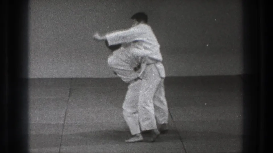 In 1971 Tokyo footage, two adult men in white gi are locked in a standing martial arts hold on a plain indoor mat.