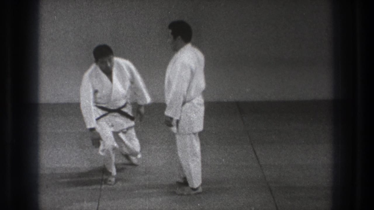 In the indoor Tokyo practice space, two men in white gi reset their stance after a martial arts floor movement.