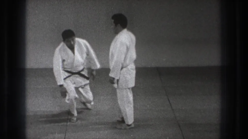 In the indoor Tokyo practice space, two men in white gi reset their stance after a martial arts floor movement.