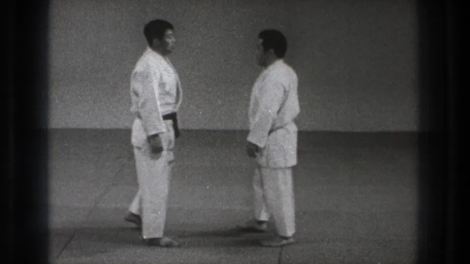 In 1971 Tokyo home-movie footage, two adult martial arts practitioners in white gi face each other before a demonstration.