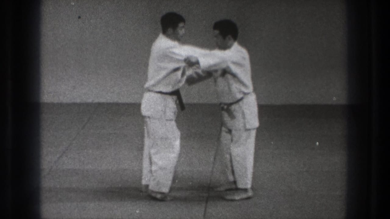 Two adult practitioners in gi continue the Harai Goshi demonstration in close contact on a plain dojo mat.