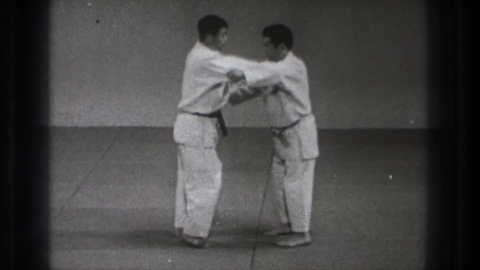 Two adult practitioners in gi continue the Harai Goshi demonstration in close contact on a plain dojo mat.