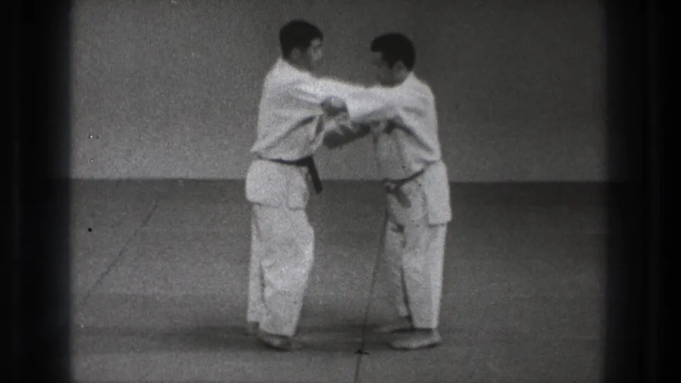 In Tokyo 1971 footage, two martial artists in white gi face each other while beginning a controlled arm-grip technique on a plain mat.