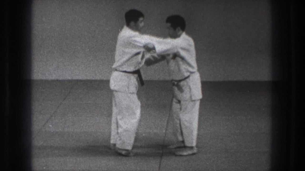 Two practitioners in white gi maintain a close arm hold during a martial arts demonstration in the Tokyo 1971 home-movie clip.