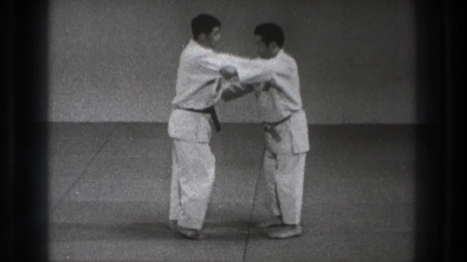 Two practitioners in white gi maintain a close arm hold during a martial arts demonstration in the Tokyo 1971 home-movie clip.