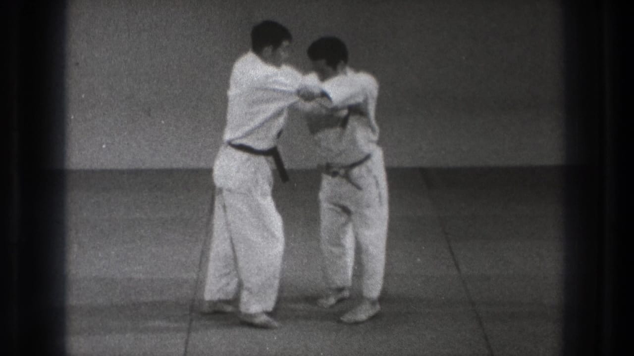 The black-belt practitioner appears to guide his partner through an arm-control movement on the mat in the 1971 Tokyo martial arts footage.