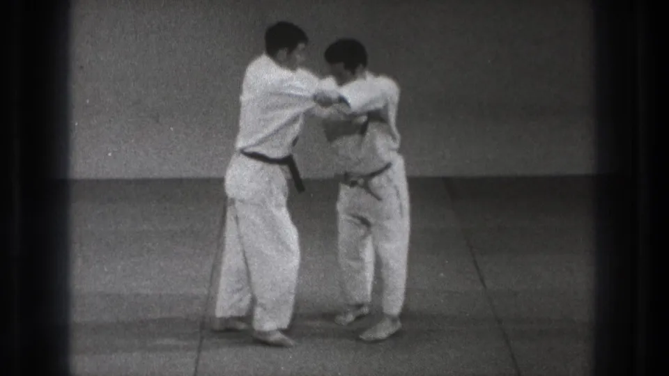 The black-belt practitioner appears to guide his partner through an arm-control movement on the mat in the 1971 Tokyo martial arts footage.