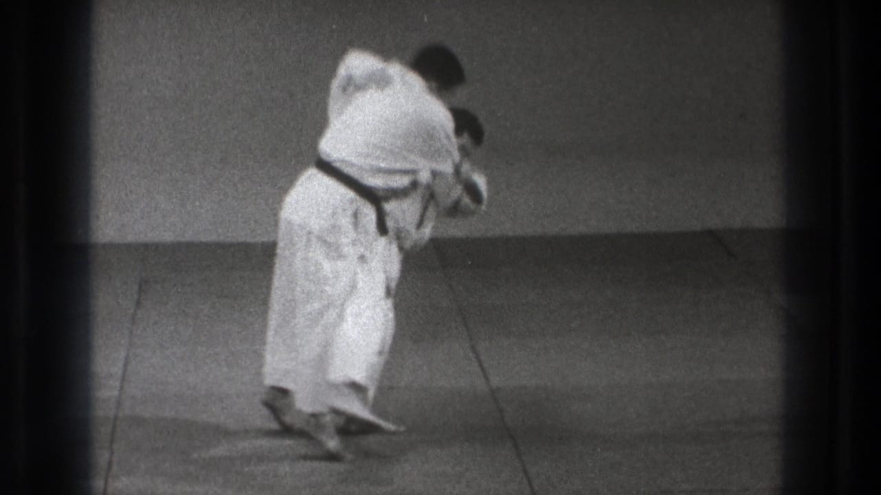 A black-belt martial artist bends his partner forward in a controlled takedown phase on the plain mat, supported by the 1971 Tokyo clip metadata.