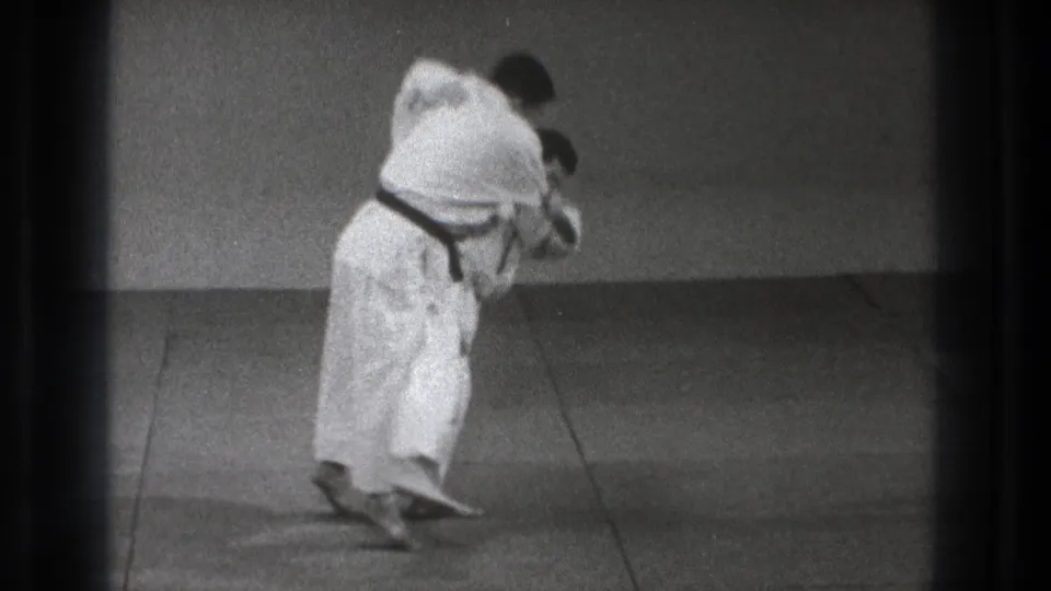 A black-belt martial artist bends his partner forward in a controlled takedown phase on the plain mat, supported by the 1971 Tokyo clip metadata.
