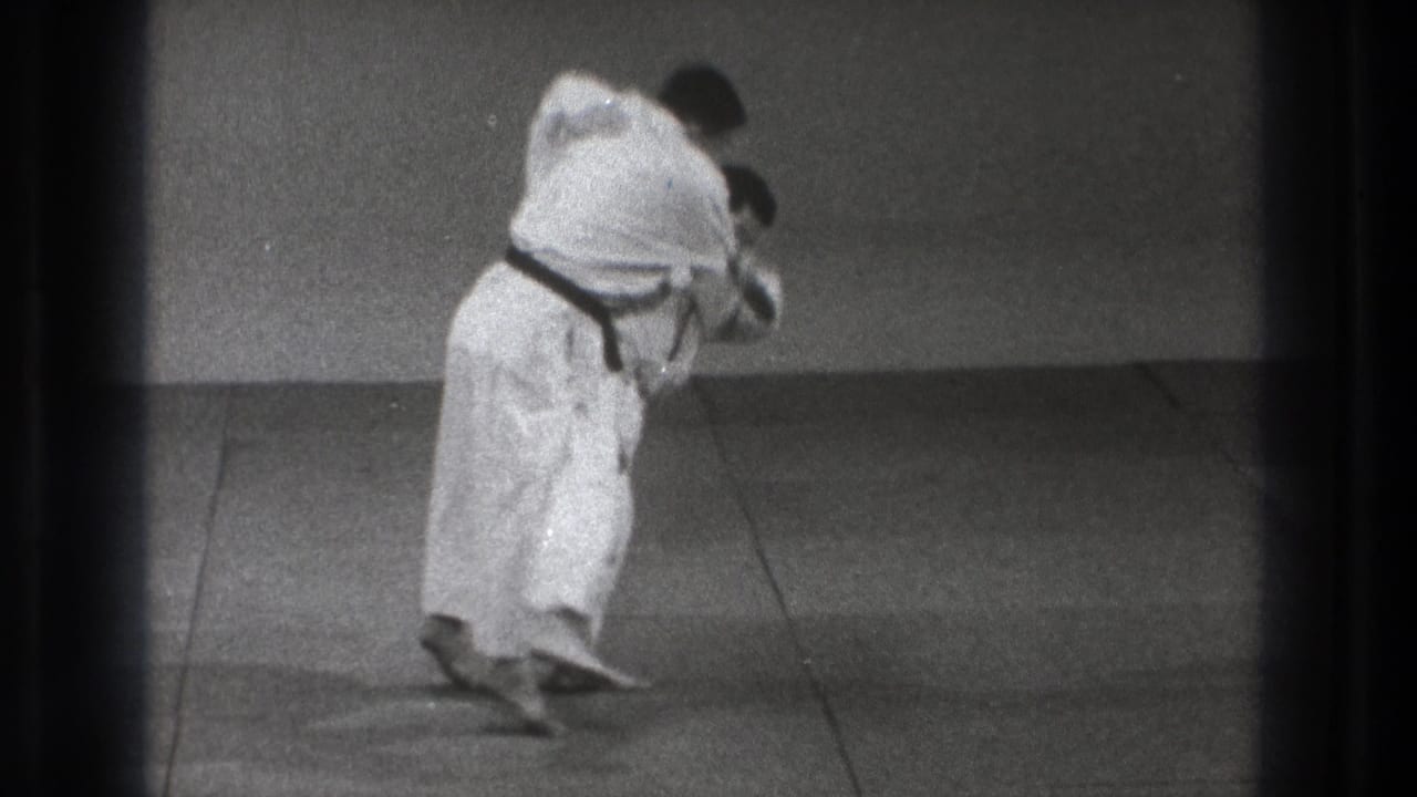 Karate practitioner in a white gi leans forward during a close hold on an indoor mat.