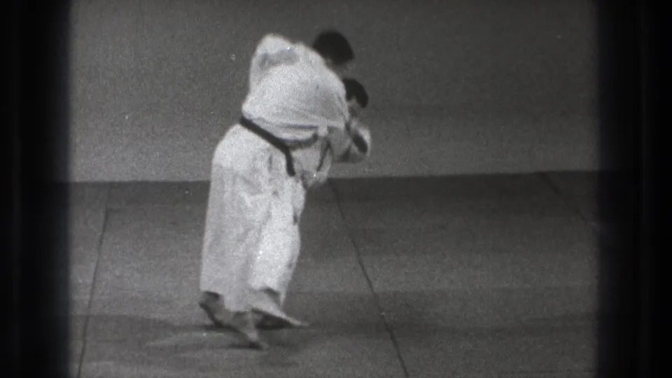 Tokyo 1971 8mm frame showing a karate practitioner in a white gi and dark belt leaning into a close balance position on a plain indoor mat.