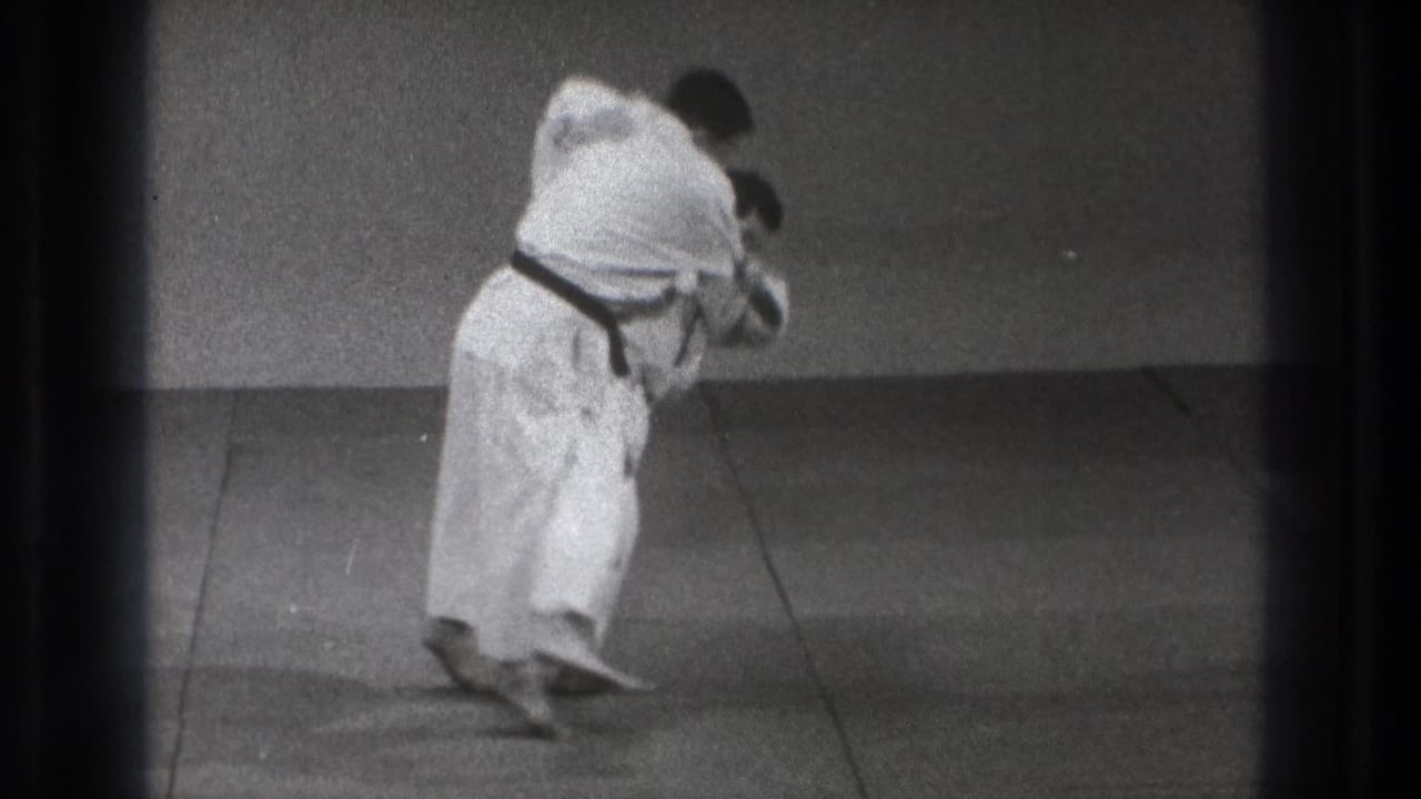 Tokyo 1971 frame of a karate practitioner in a white gi and dark belt maintaining a forward-leaning stance on an indoor practice mat.