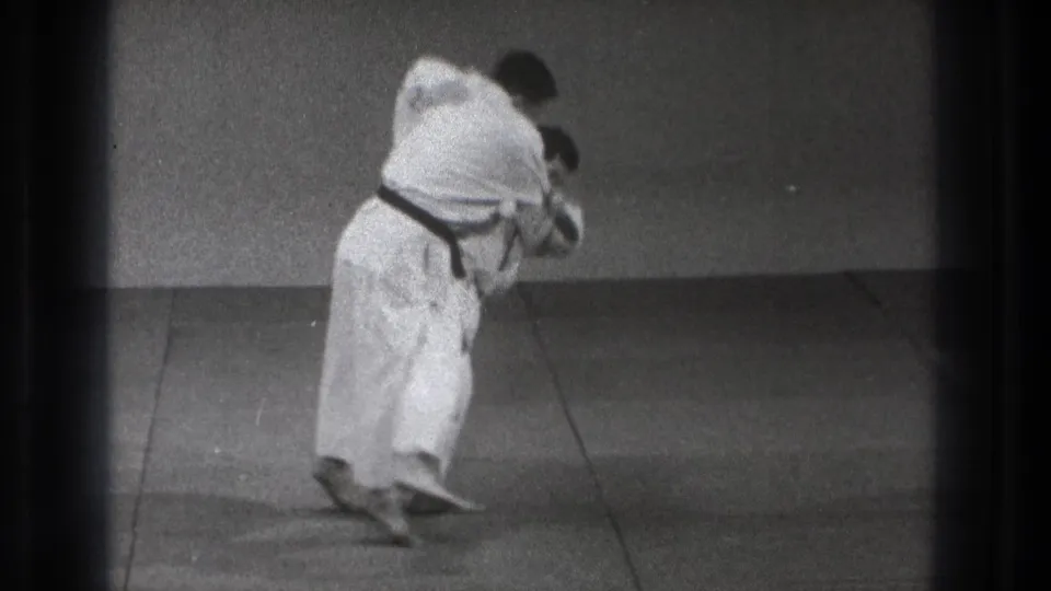 Tokyo 1971 frame of a karate practitioner in a white gi and dark belt maintaining a forward-leaning stance on an indoor practice mat.