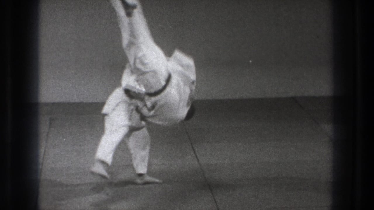 Tokyo 1971 8mm frame showing a karate practitioner in mid-throw, one leg raised high as the body rotates above an indoor mat.