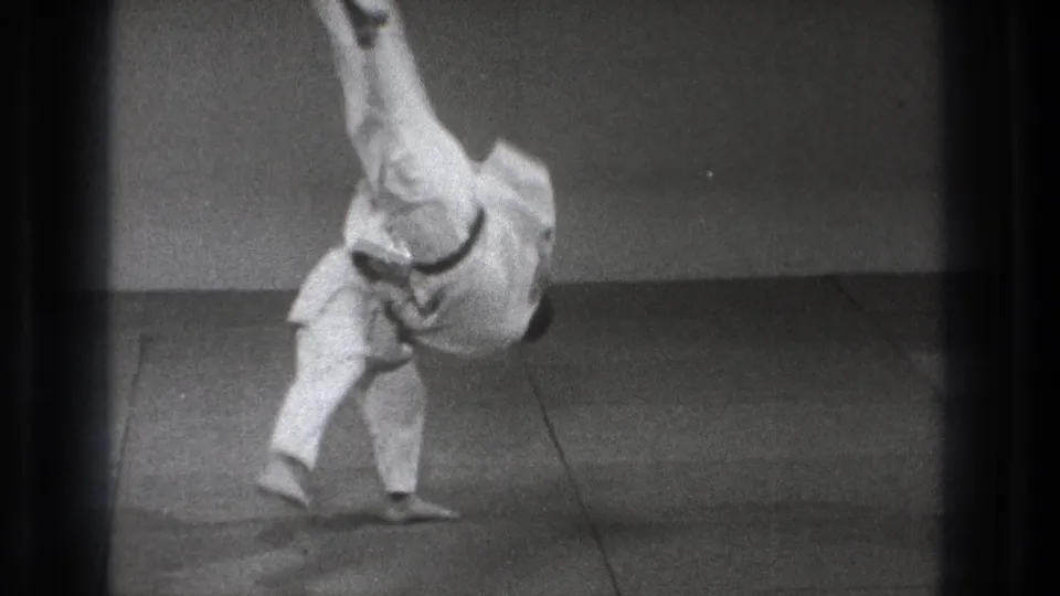 Tokyo 1971 8mm frame showing a karate practitioner in mid-throw, one leg raised high as the body rotates above an indoor mat.