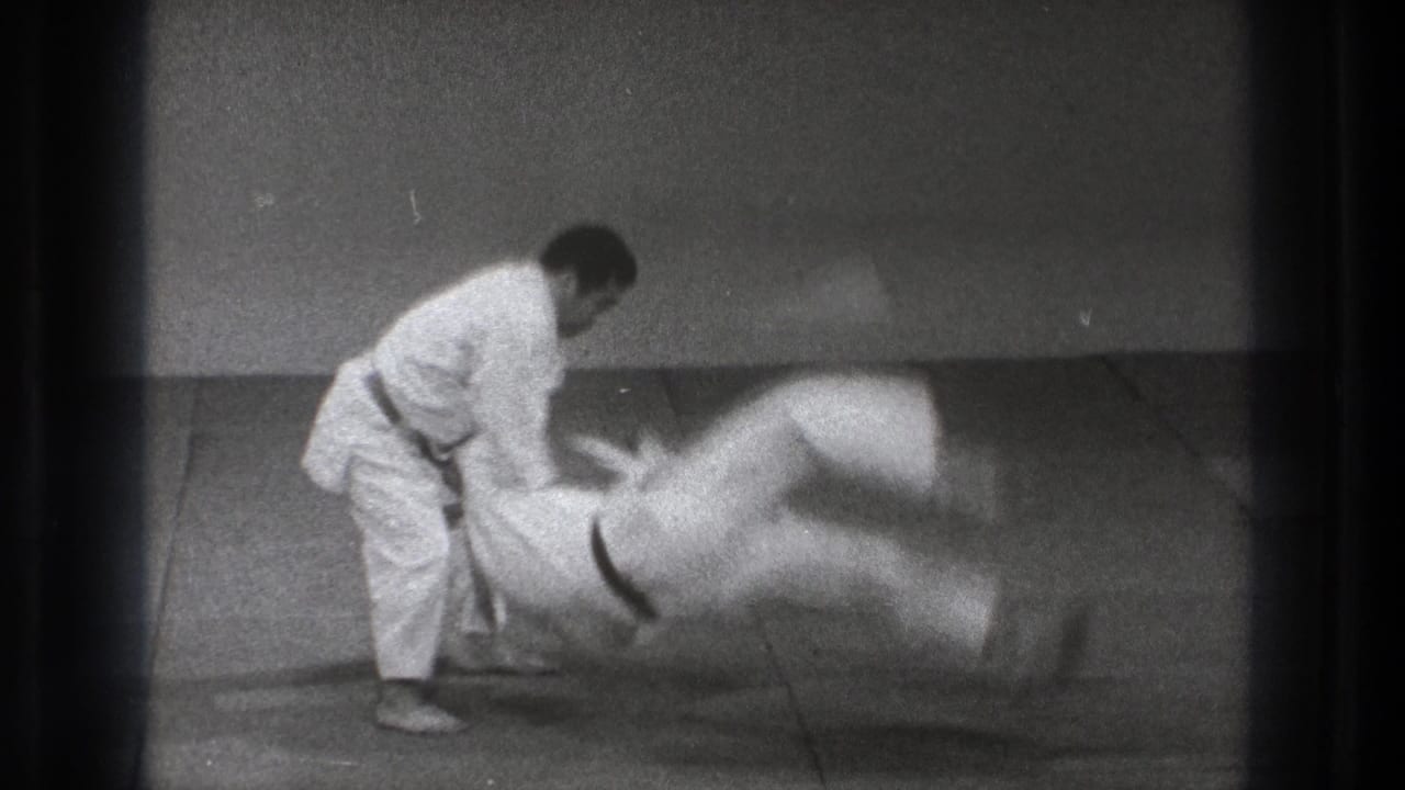 Two men in white gi practice a throw on a mat in a plain dojo space.