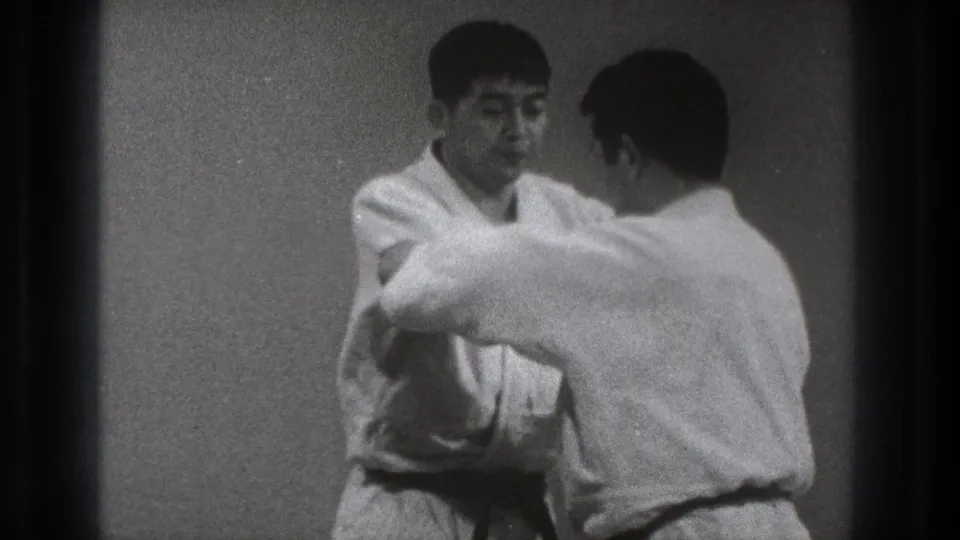 Two adult men in white gi practice close-quarters karate technique in a plain Tokyo dojo setting, based on supplied 1971 metadata.
